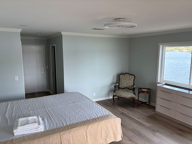 Bedroom with light blue walls, wood flooring, bed, chair, dresser, and window overlooking water.