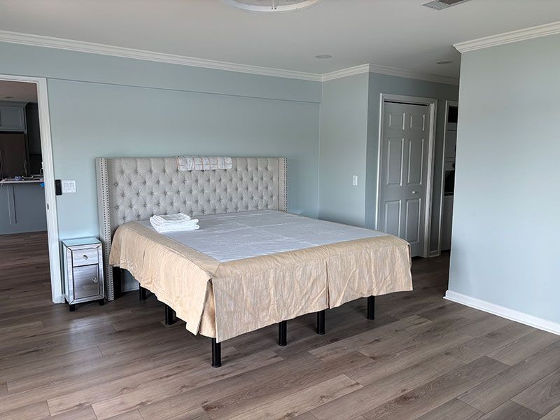 Bedroom with a bed, nightstand, and light blue walls. Wooden floors and a white door are visible.