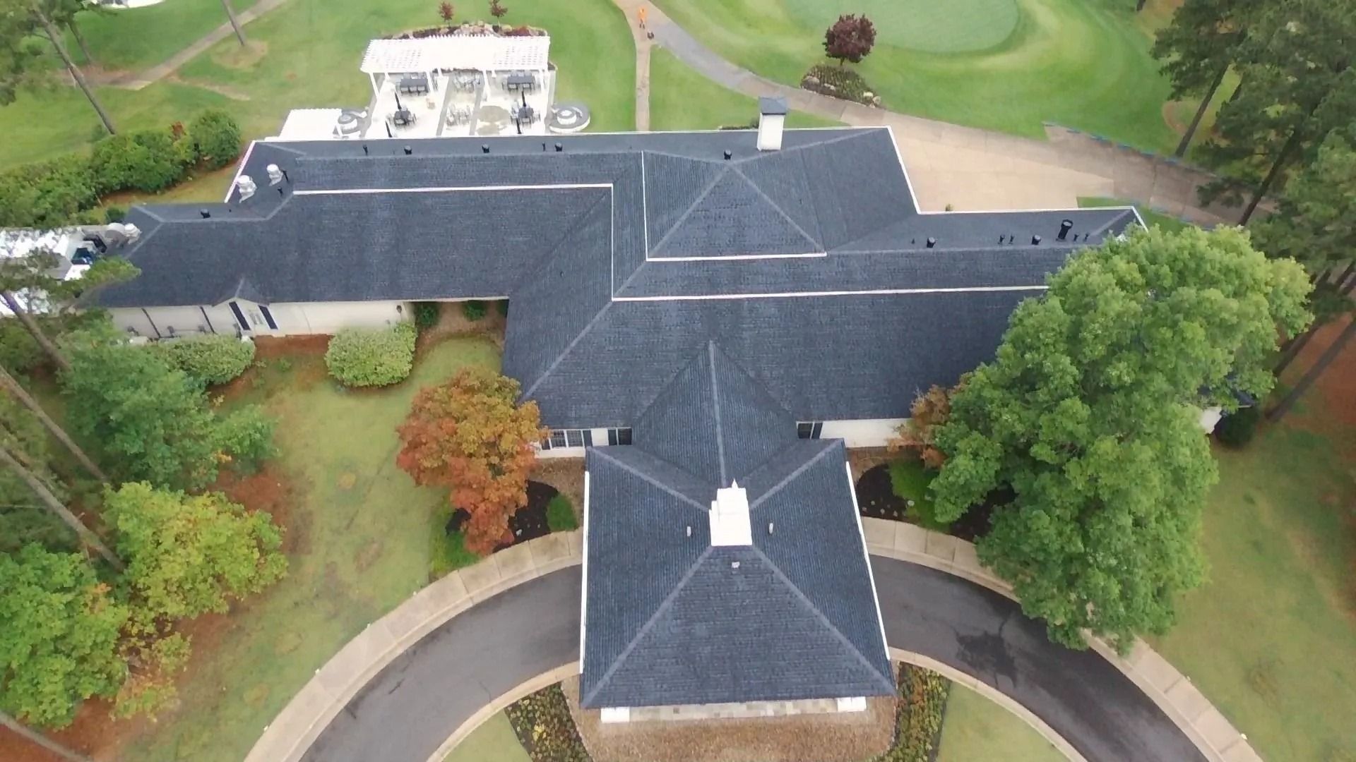 Overhead view of a large, dark-roofed house on a golf course with a circular driveway and surrounding trees.