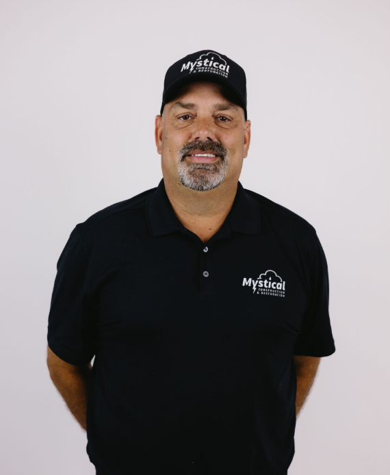 Man in black polo and cap standing against a plain light background, smiling.