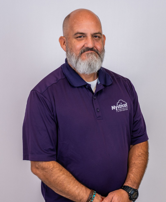 Man in a purple polo shirt with a white beard, standing against a light gray background.