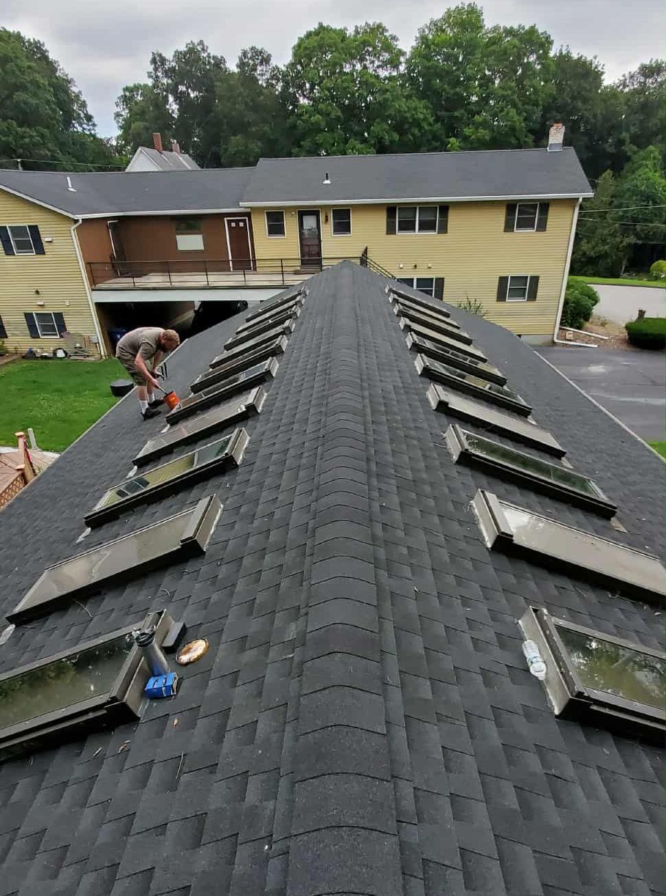 Roofers installing skylights on a dark shingled apartment roof, with buildings and trees in the background.