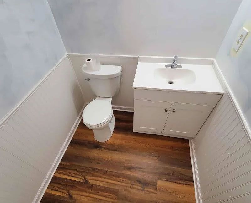 Small bathroom with white toilet and sink vanity, beadboard walls, and wood-look floor.