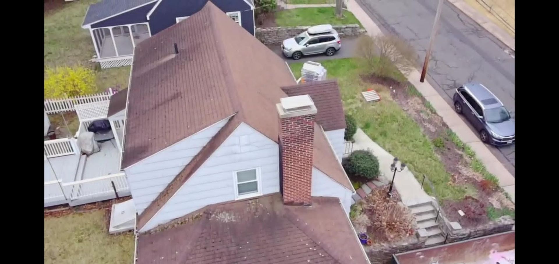 Aerial view of a house with a brown roof, brick chimney, and parked cars in the driveway and street
