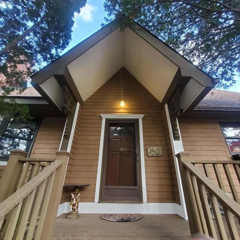 Front porch entrance of a tan house with wood siding, peaked awning, steps, and a lit lamp above the door