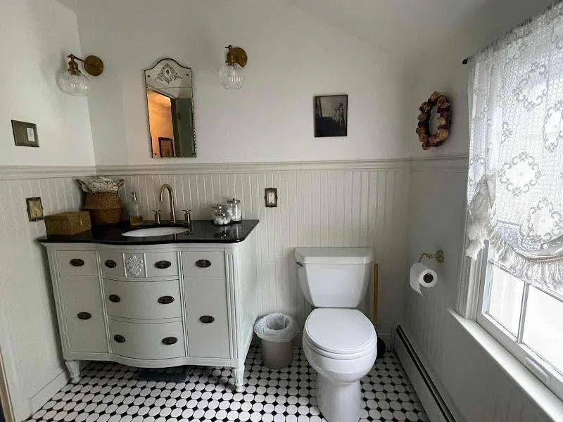 Small bathroom with white walls, pedestal sink, toilet, wicker basket, and window with lace curtain.