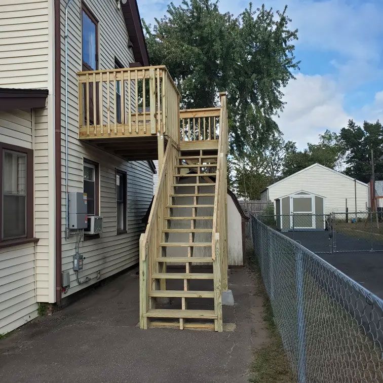 Wooden outdoor stairs and balcony attached to a house beside a chain-link fence and driveway