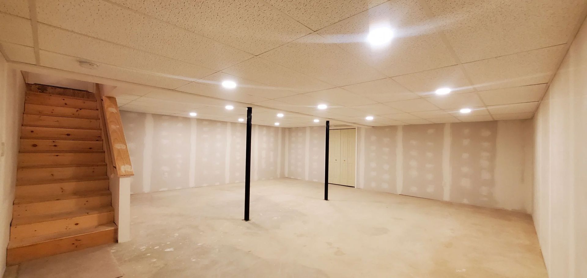 Empty unfinished basement with concrete floor, wooden stairs on left, and support poles in the center.