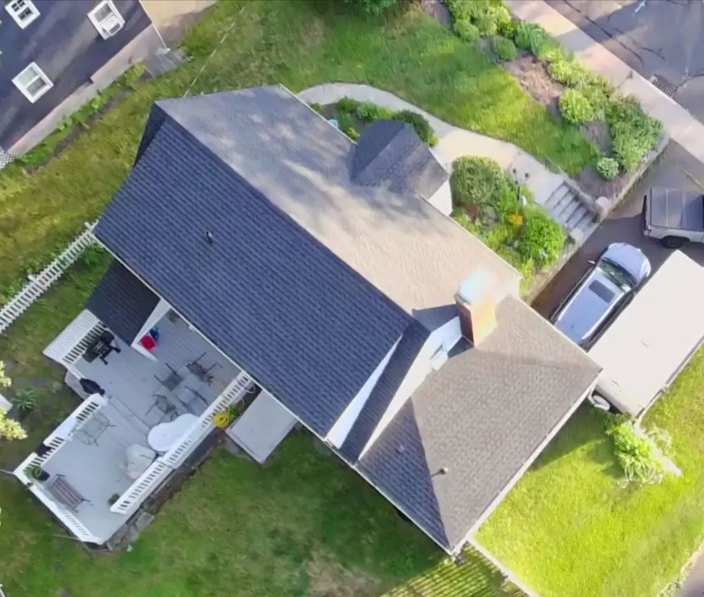 Aerial view of a house with a dark roof, driveway, green lawn, and parked cars.