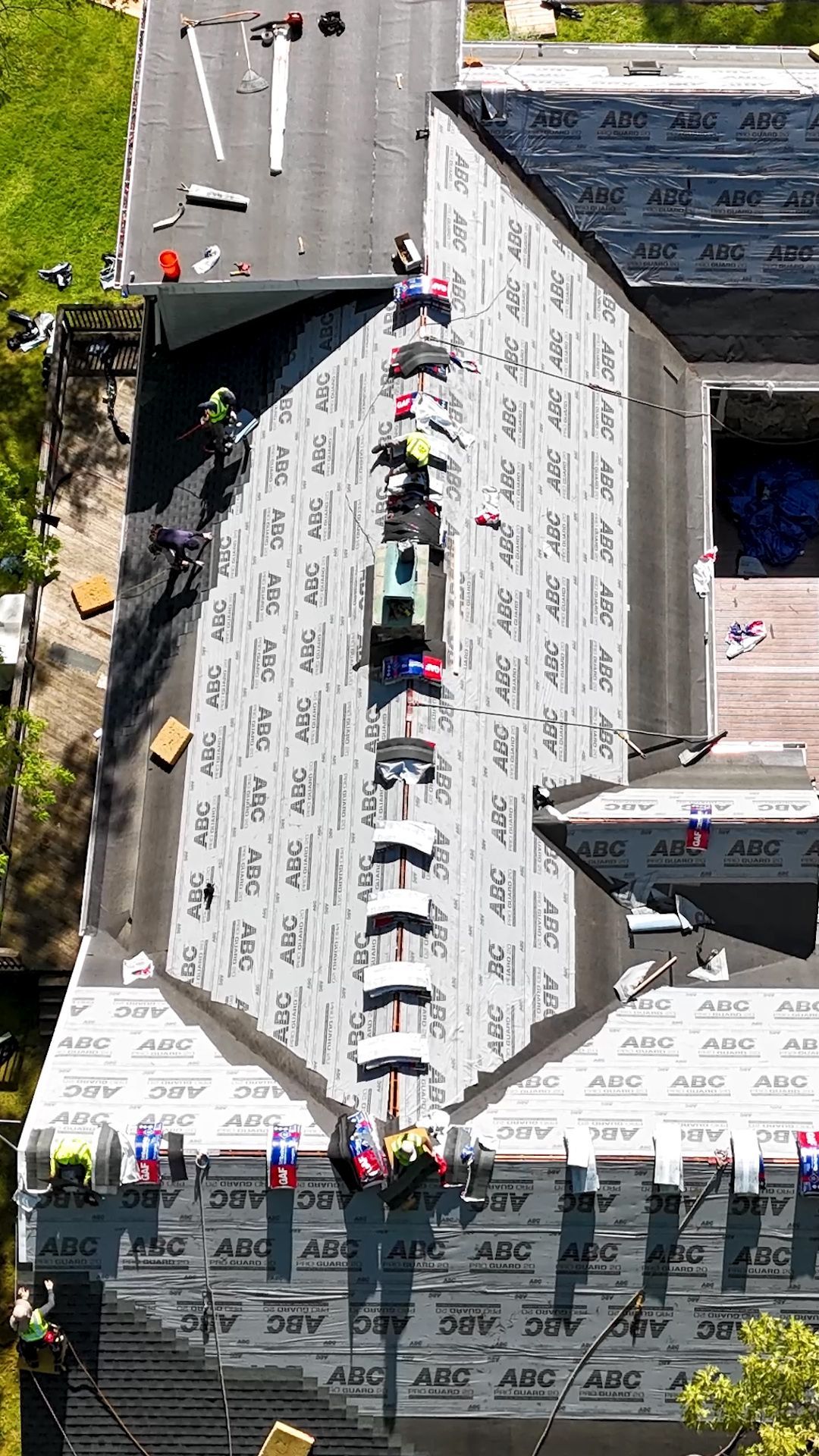 Aerial view of workers on a roof, wearing safety gear and standing along a central ridge near parked trucks.