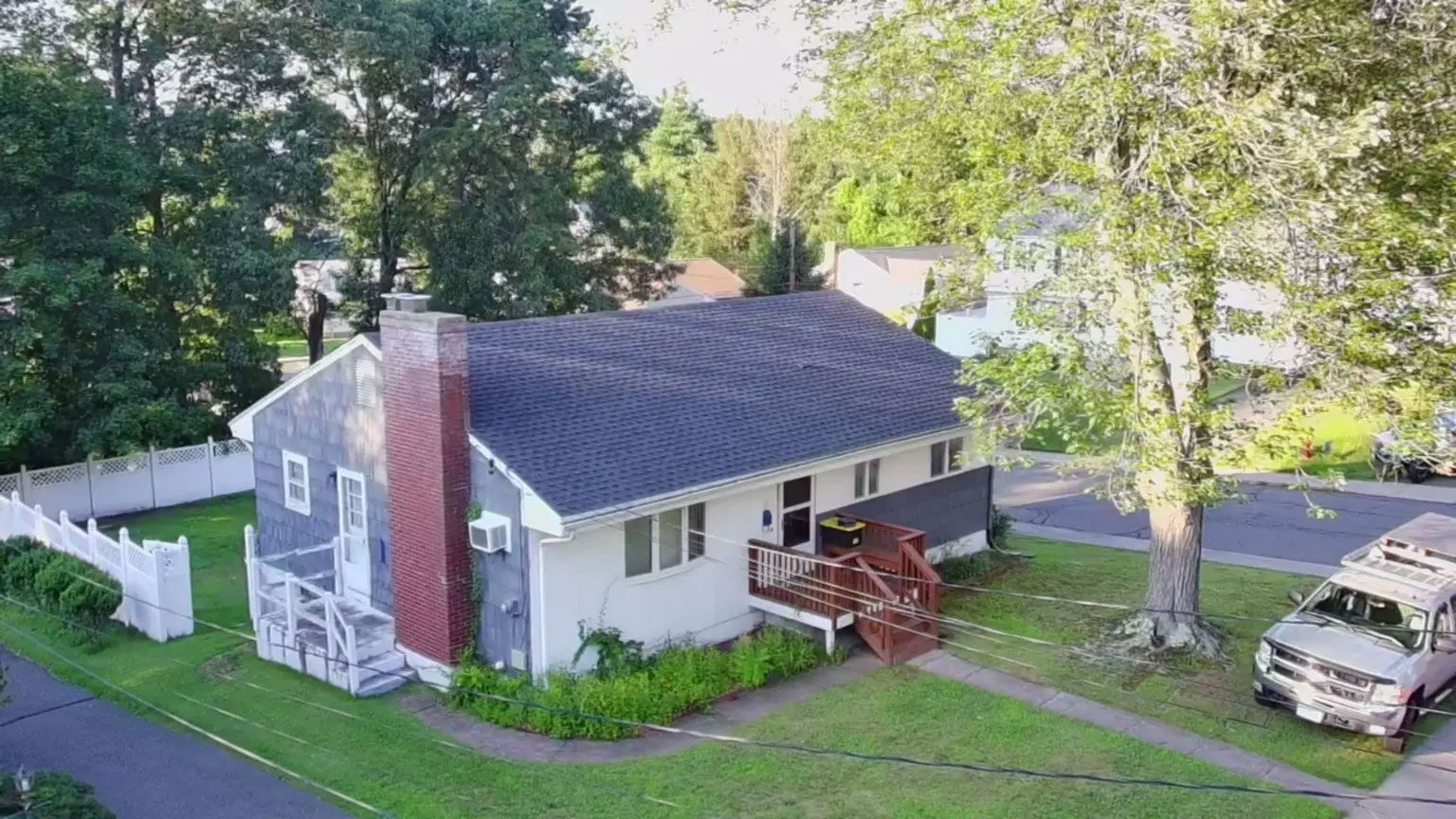 Small white house with a red brick chimney, a wooden porch, and a car parked beside a tree-lined yard.