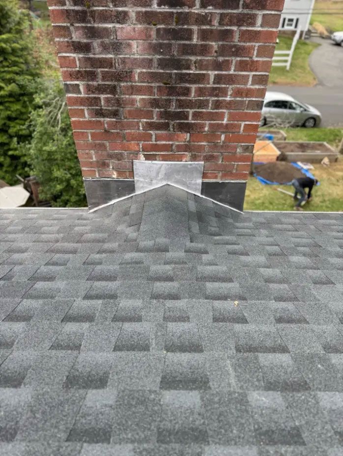 Rooftop shingles lead to a brick chimney with metal flashing; street and parked car below.