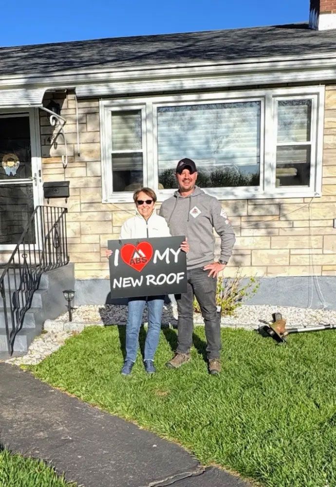 Two people holding a sign that says “I ♥ MY NEW ROOF” in front of a house
