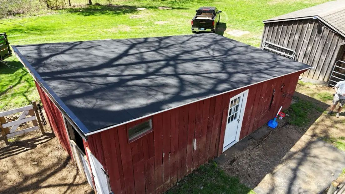 Red barn with dark roof in a grassy yard, with a vehicle parked behind it and a wooden deck nearby.