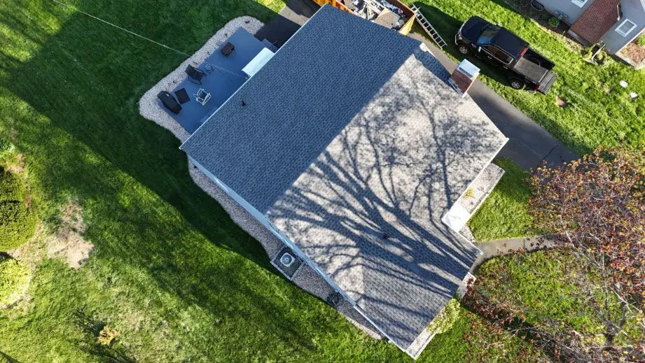 Aerial view of a house with a gray roof, green lawn, and tree shadows