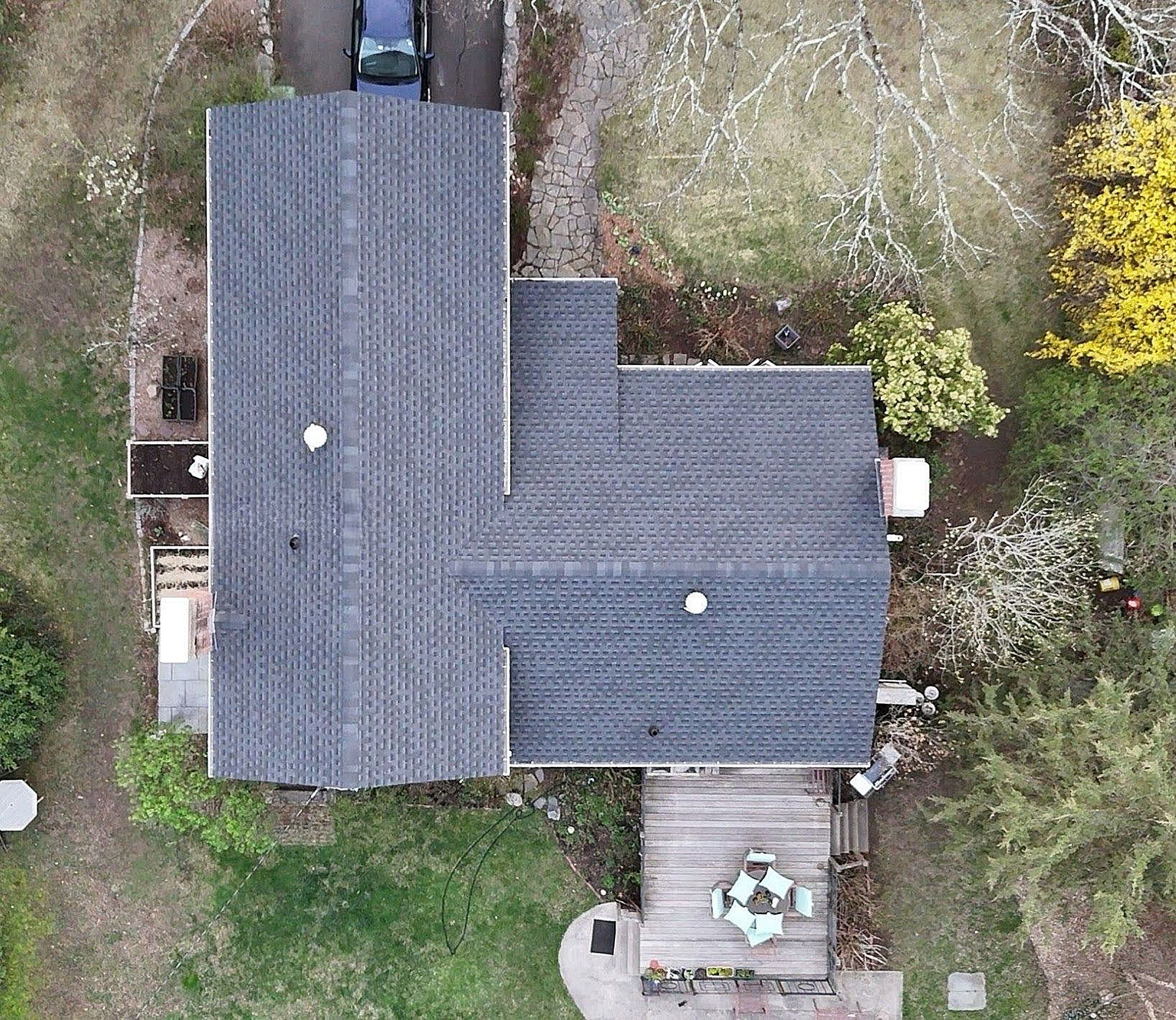 Aerial view of a gray-roofed house with a small backyard patio and surrounding trees.