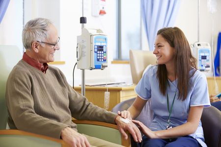 Man receiving IV treatment; nurse in blue scrubs smiles and holds his hand in a hospital room.