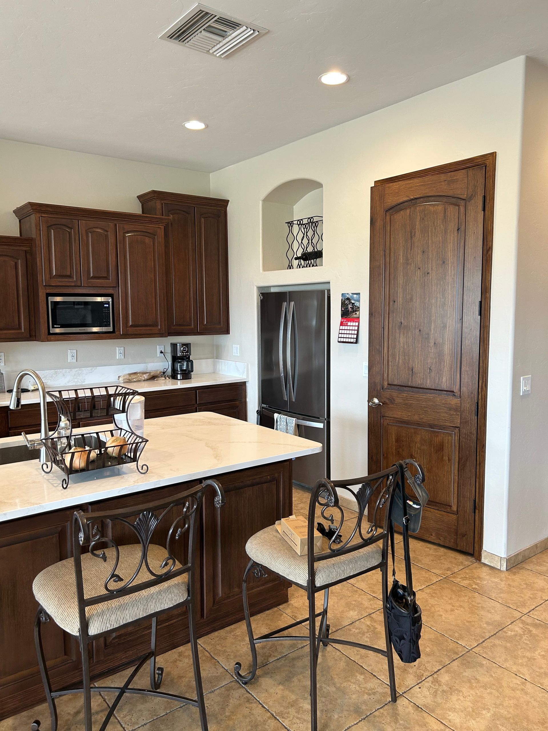 Kitchen with dark wood cabinets, white island, stainless fridge, and wrought-iron stools on tiled floor.