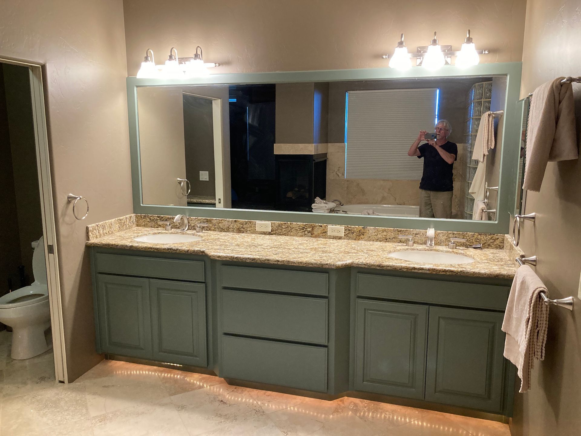 Bathroom vanity with green cabinets, granite countertop, large mirror, and wall lights; person visible in mirror.