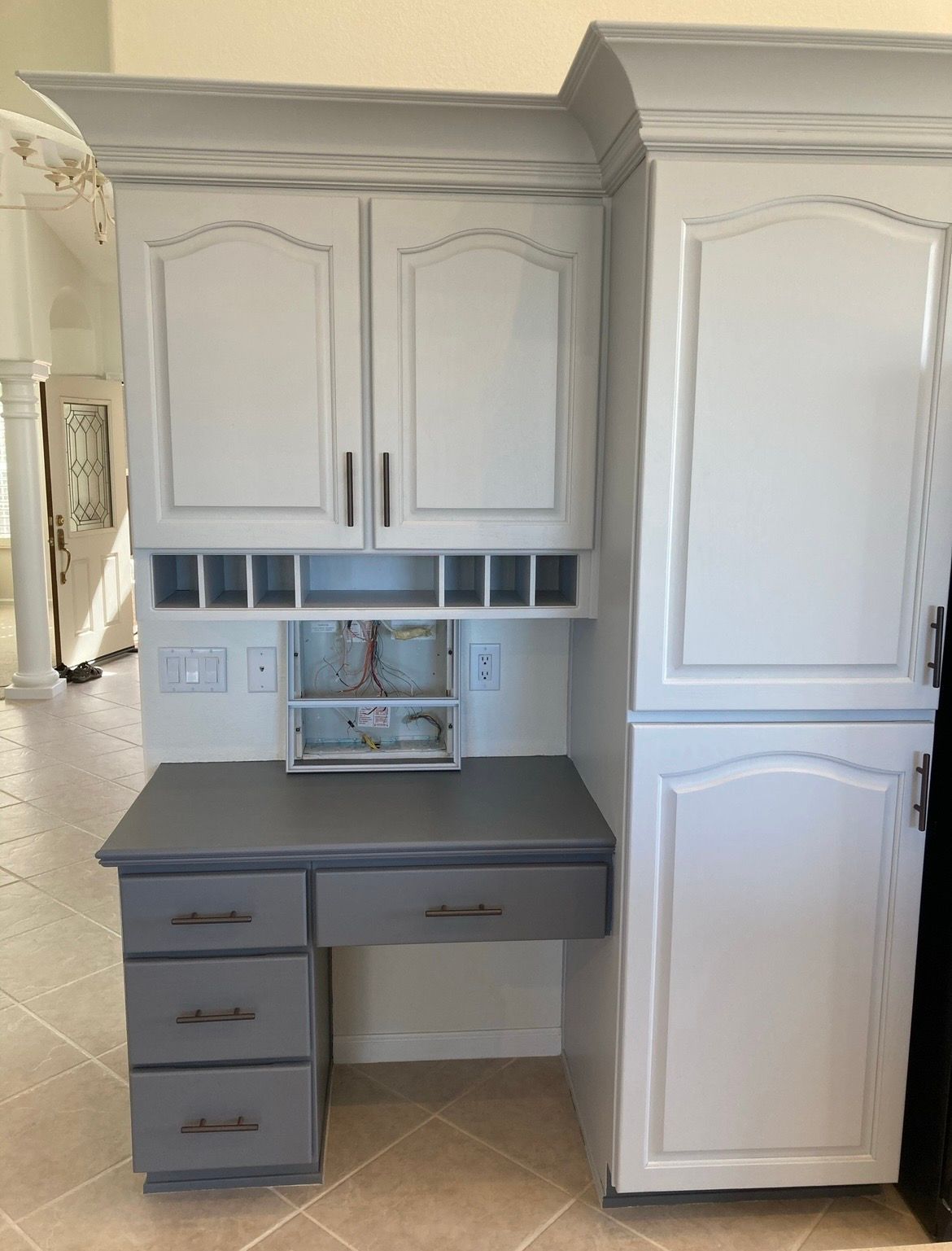 Gray built-in desk nook with cabinets, drawers, and white pantry cabinets in a tiled kitchen area.