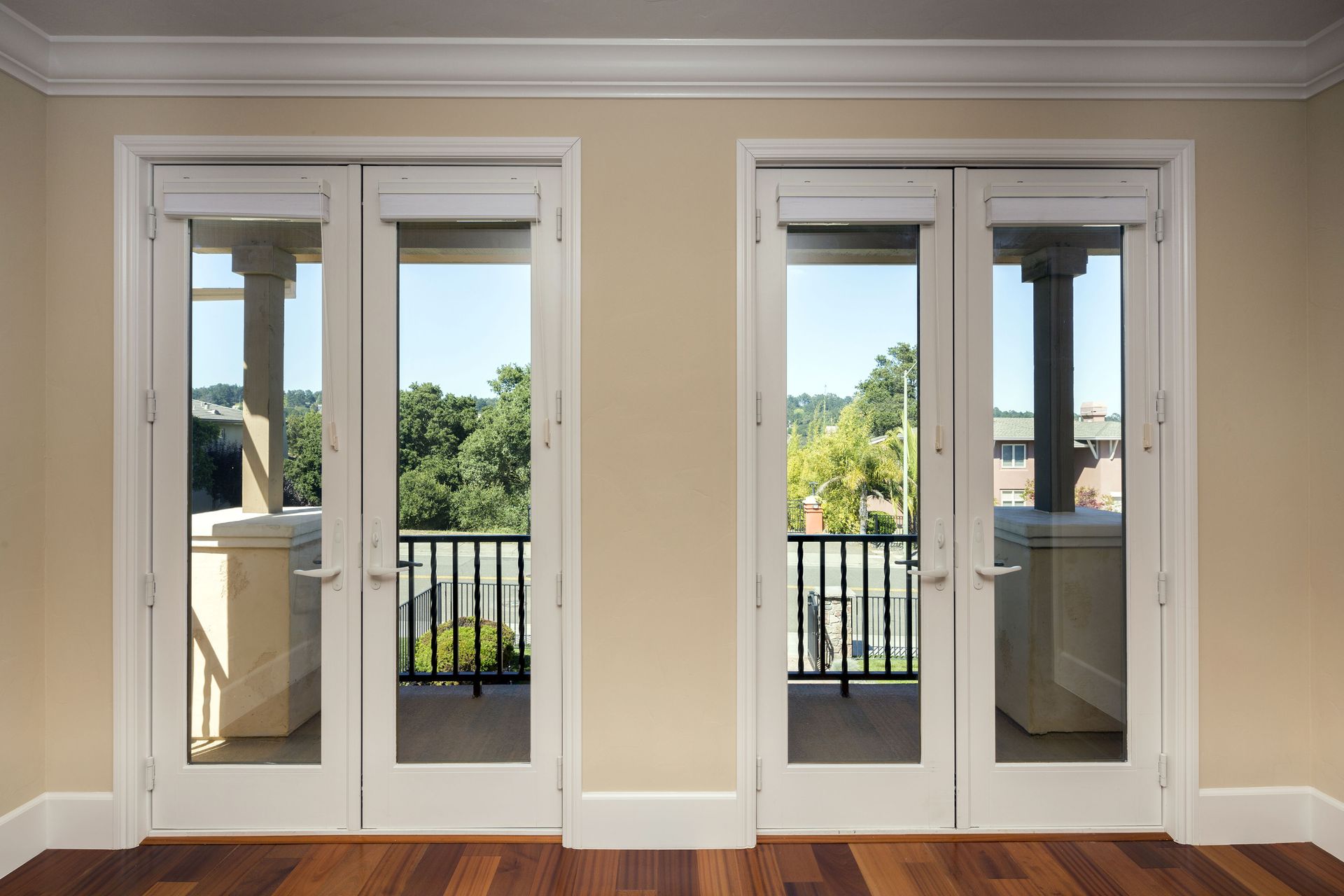 Two white french doors with glass panels open to a balcony with trees, a beige wall, and hardwood floor.