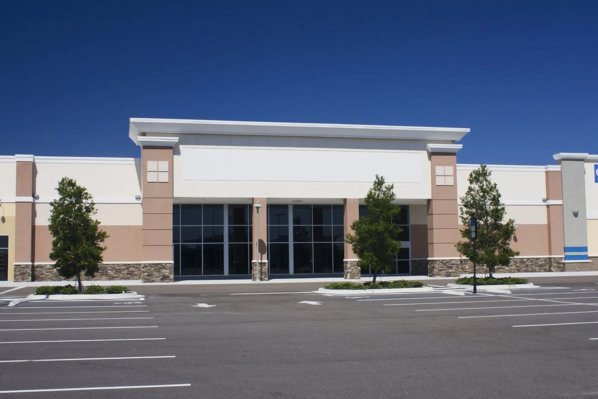 Exterior of a vacant commercial building with large windows and a mostly empty parking lot under a blue sky.