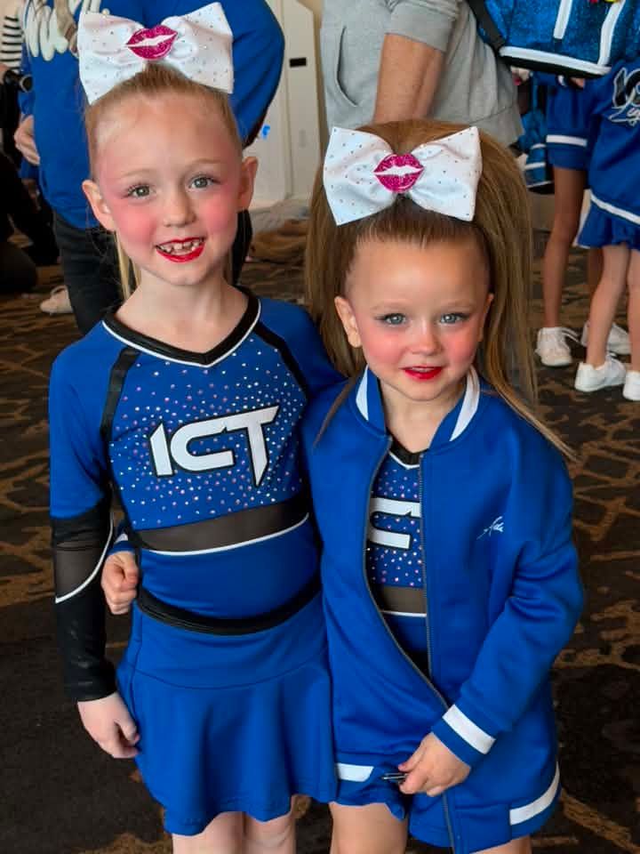 Two children in blue cheer uniforms with white bows and red lipstick standing together indoors.
