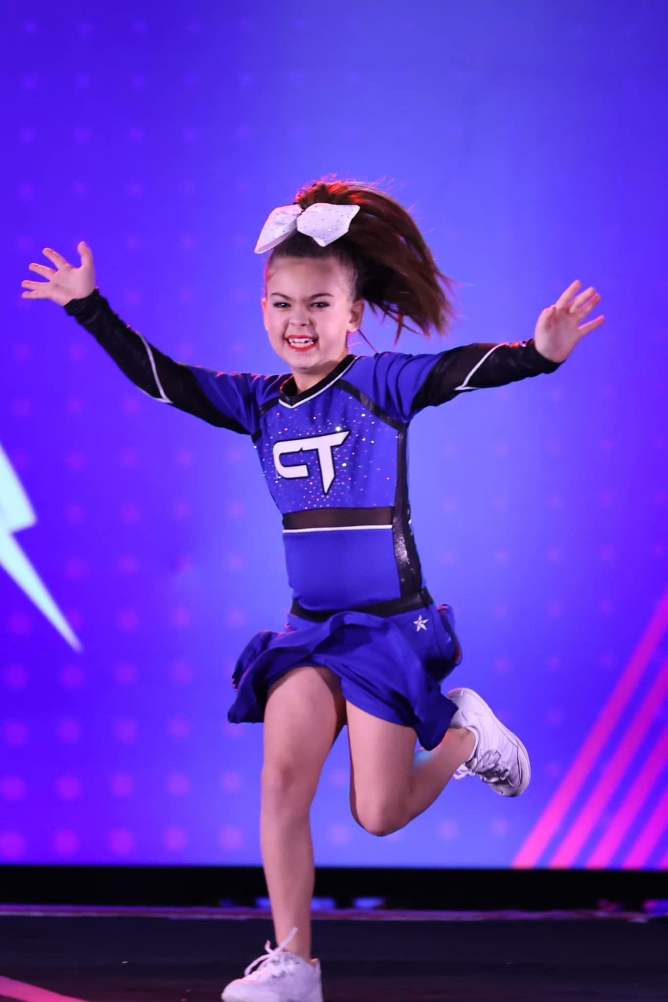 A young girl in a blue cheerleading uniform is running on a stage.