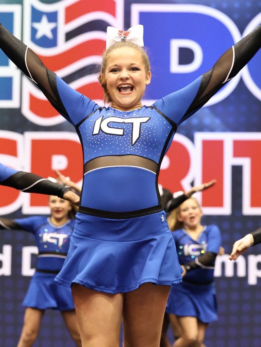 Cheerleader in blue uniform with arms raised, smiling at competition.