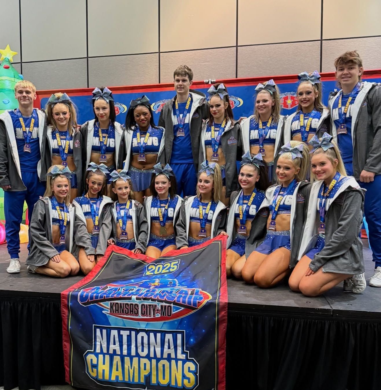 Cheerleading team posing with a banner. They wear blue & white uniforms and medals. They are on a stage.