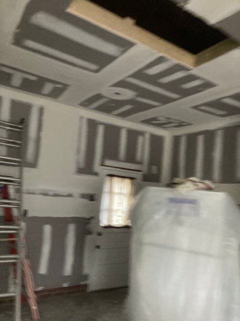 Interior view of a room under construction, drywall on walls and ceiling, ladder, door, and plastic-wrapped materials.