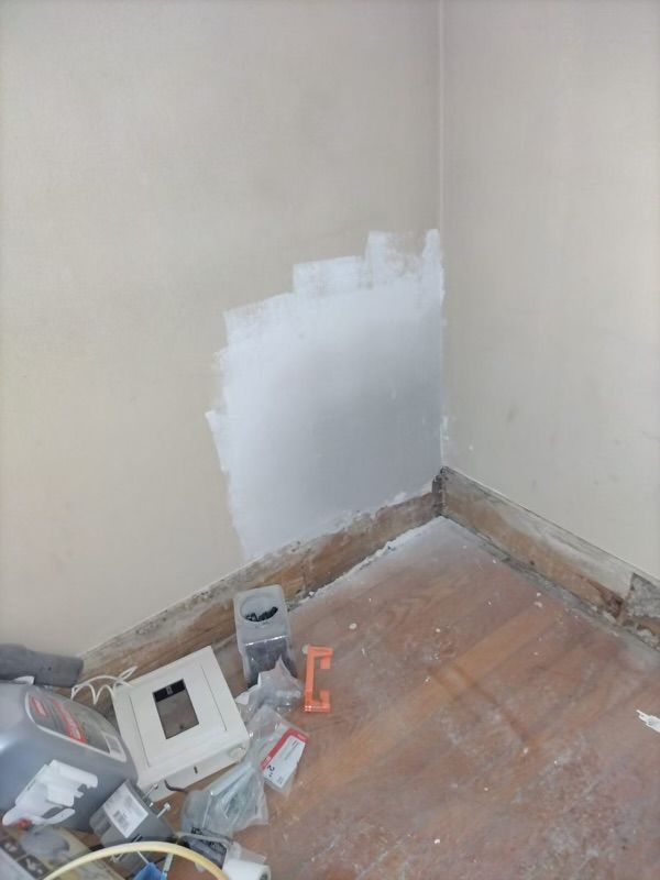 Corner of a room under renovation with unfinished walls and wooden floor covered in debris.