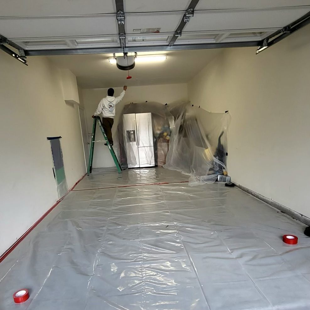 Person painting garage ceiling, standing on ladder. Walls are white, floor covered with plastic sheeting.