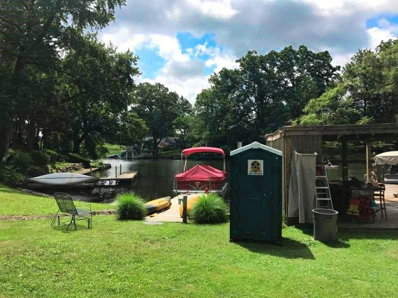 A green portable toilet is sitting in the grass next to a boat.