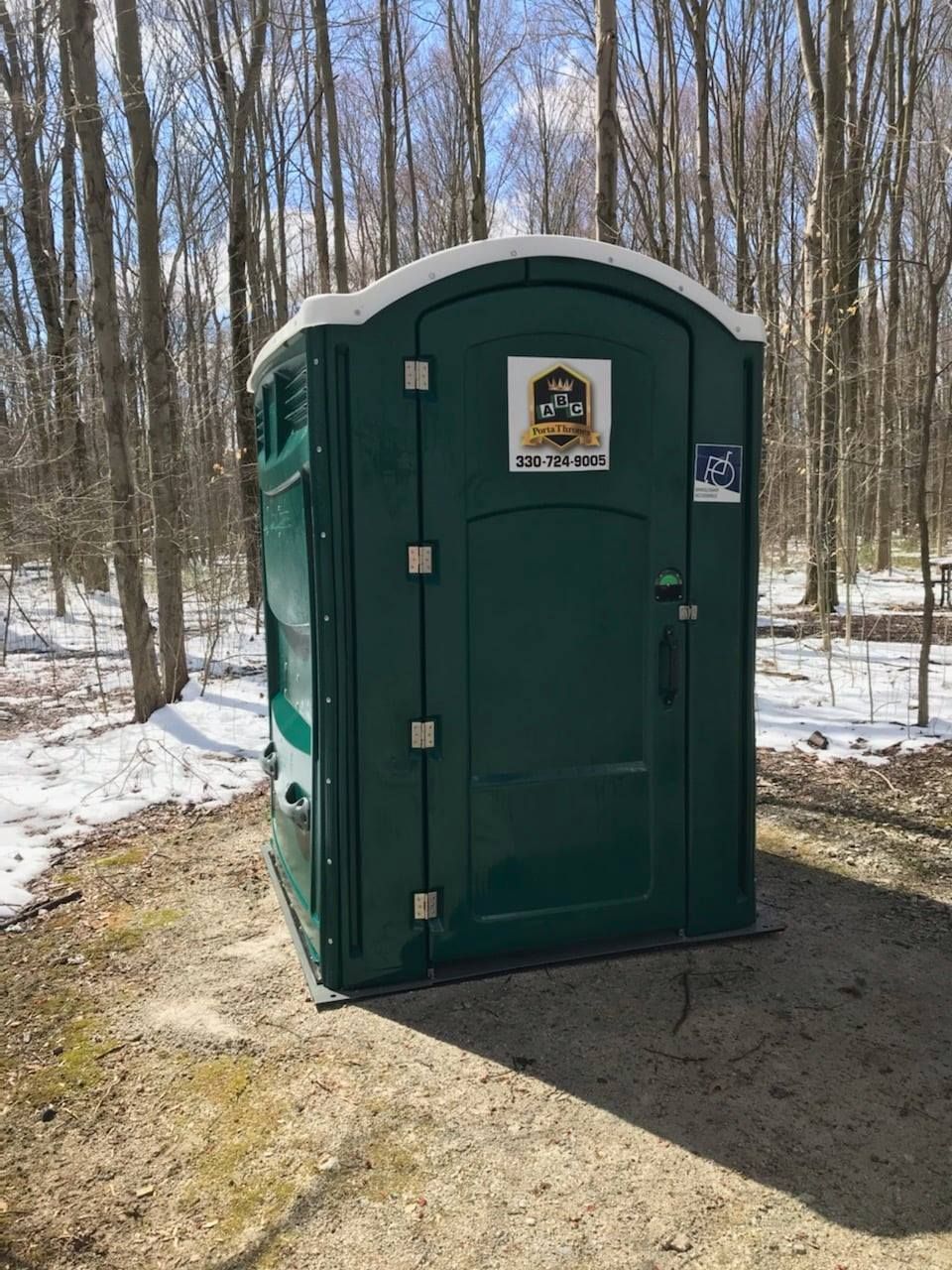 A green portable toilet is sitting in the middle of a snowy forest.