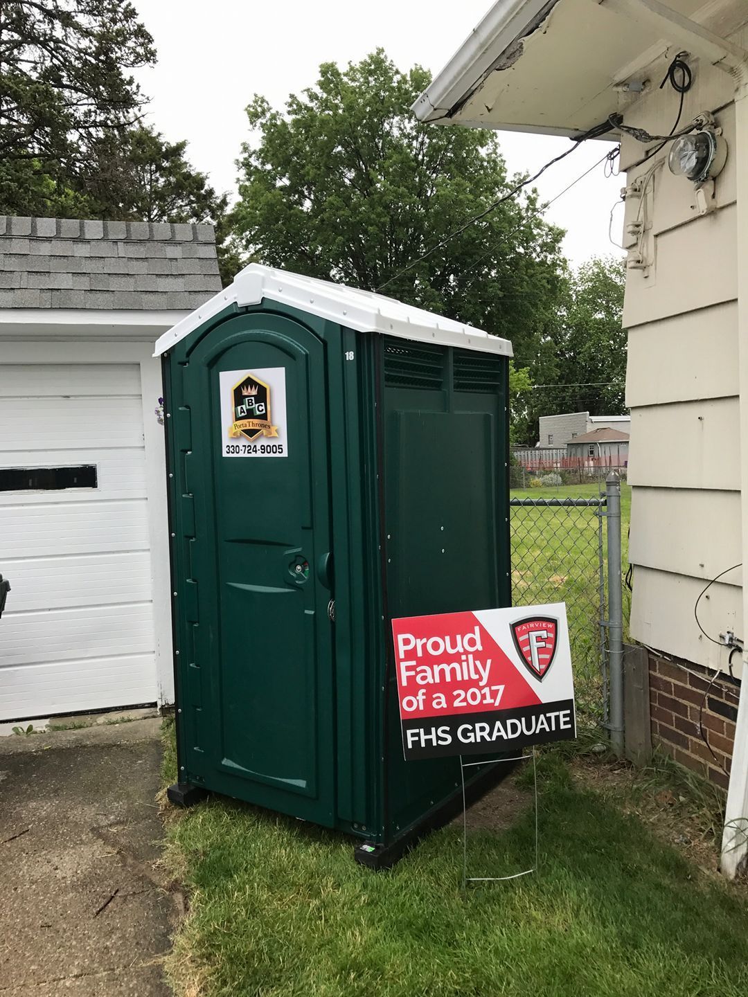 A green portable toilet is sitting in the grass next to a house.
