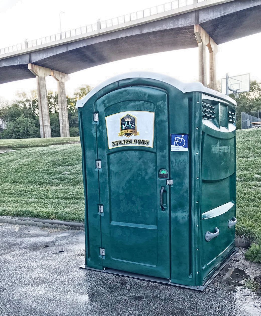 A green portable toilet is sitting in front of a bridge.