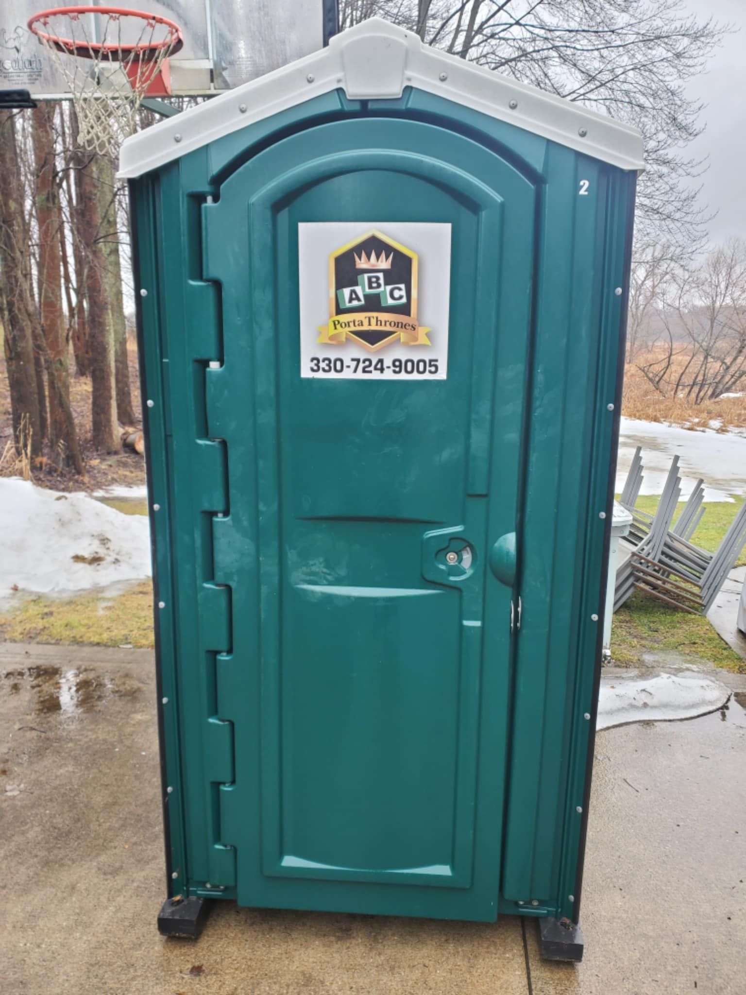 A green portable toilet is sitting on a sidewalk next to a basketball hoop.