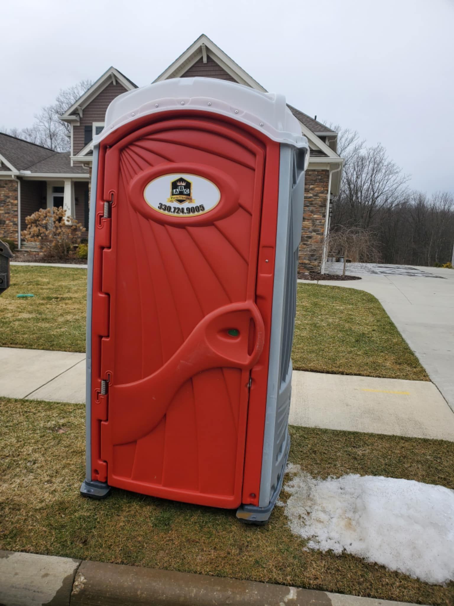 A red portable toilet is sitting in front of a house covered in snow.