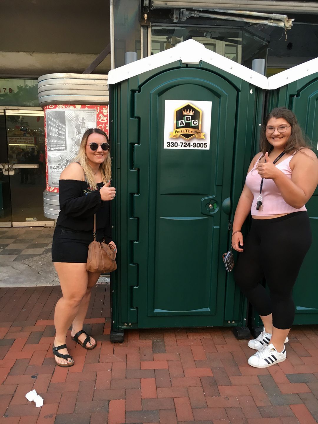 Two women are standing next to a green portable toilet on a brick sidewalk.