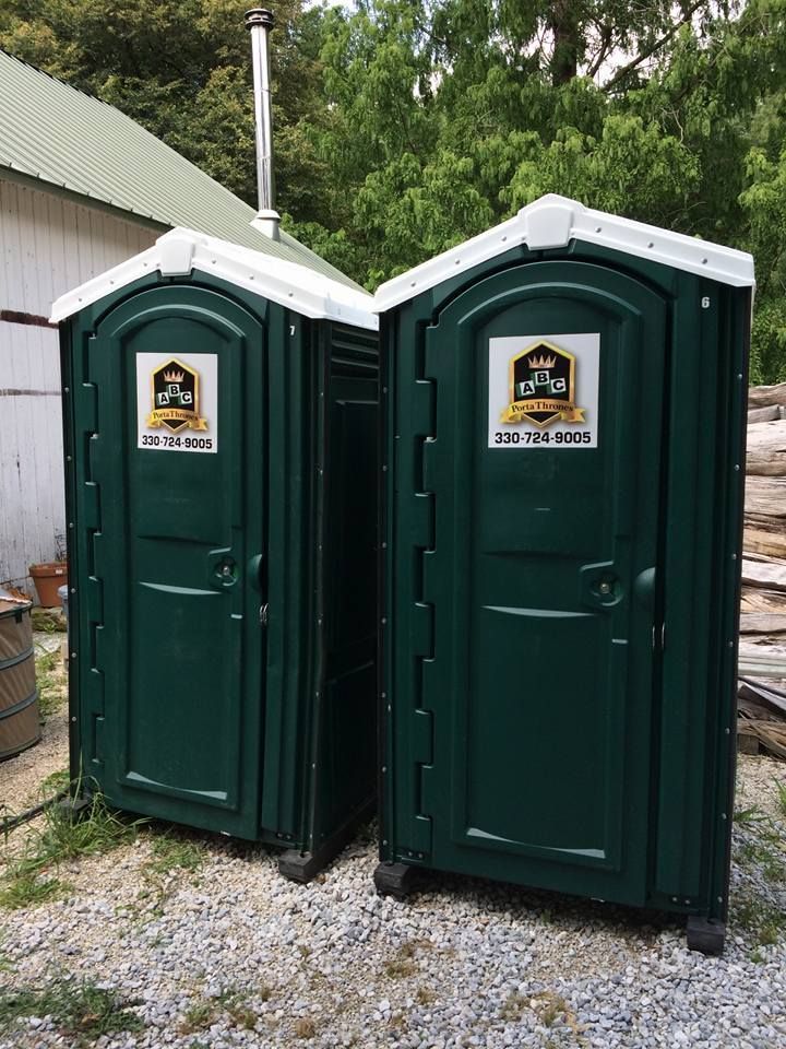 Two green portable toilets are sitting next to each other on gravel in front of a building.
