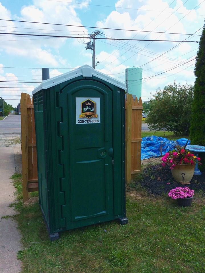 A green portable toilet is sitting in the grass next to a wooden fence.