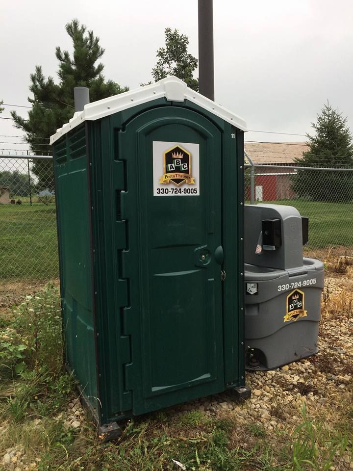 A green portable toilet is sitting in the grass next to a trash can.