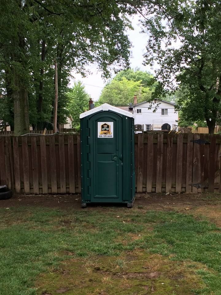 A green portable toilet is sitting in the grass in front of a wooden fence.