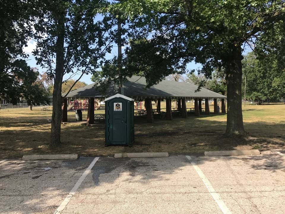 A green portable toilet is in a parking lot in front of a pavilion.