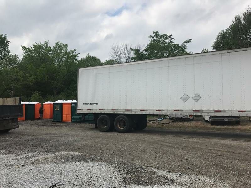 A white trailer is parked in a gravel lot next to a row of portable toilets.