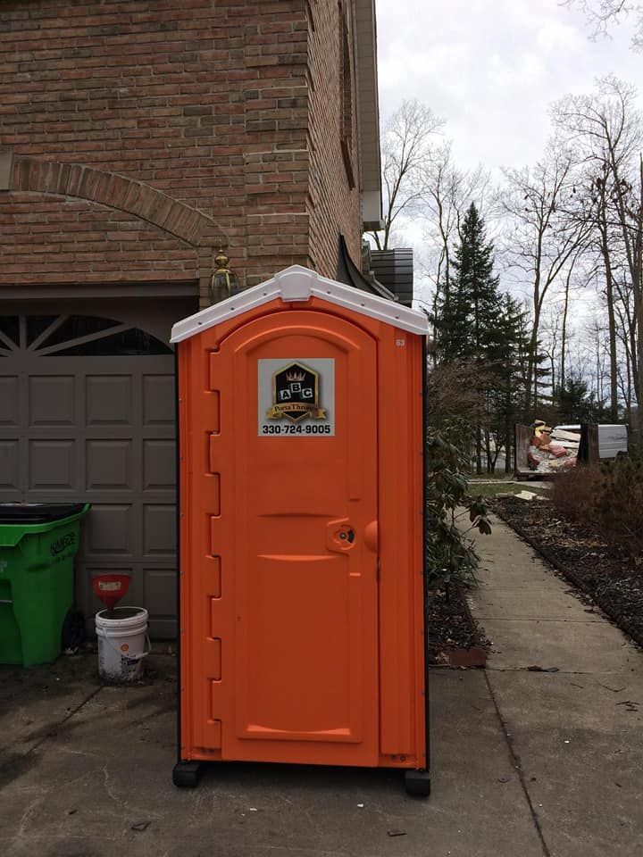 An orange portable toilet is parked on the sidewalk in front of a brick building.