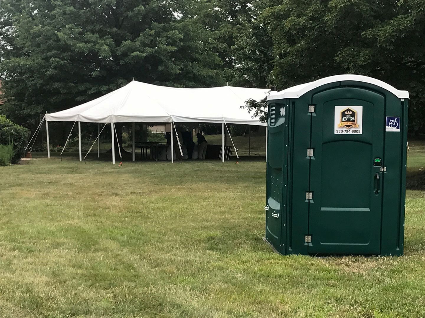 A green portable toilet is sitting in a grassy field next to a white tent.