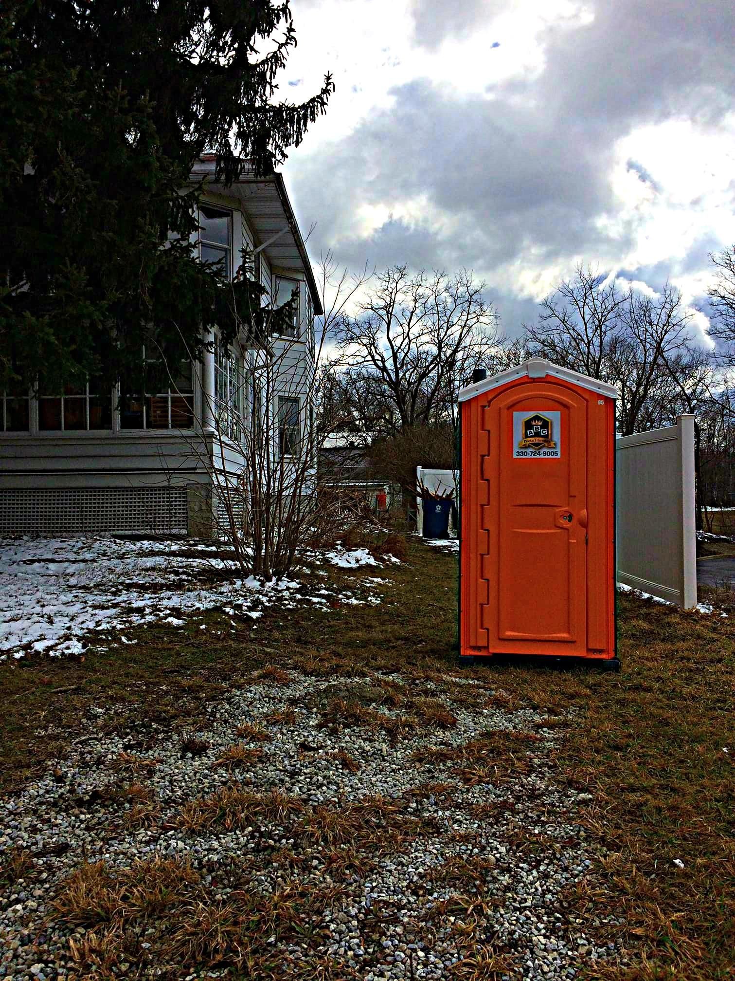 An orange portable toilet is sitting in the grass in front of a house.