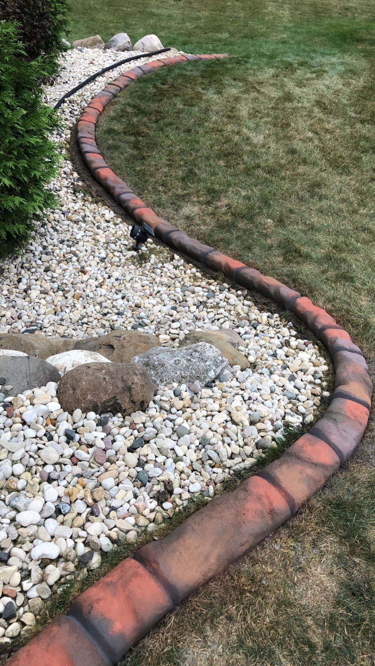 A brick curb is surrounded by rocks and grass in a yard.